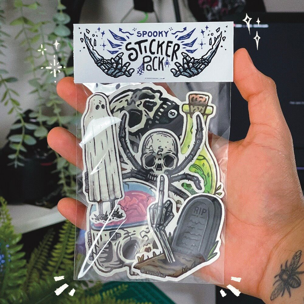 En hand som håller ett sticker pack med tecknade klistermärken i spöktema och en header där det står ”SPOOKY STICKER PACK”, mot en bakgrund av växter
