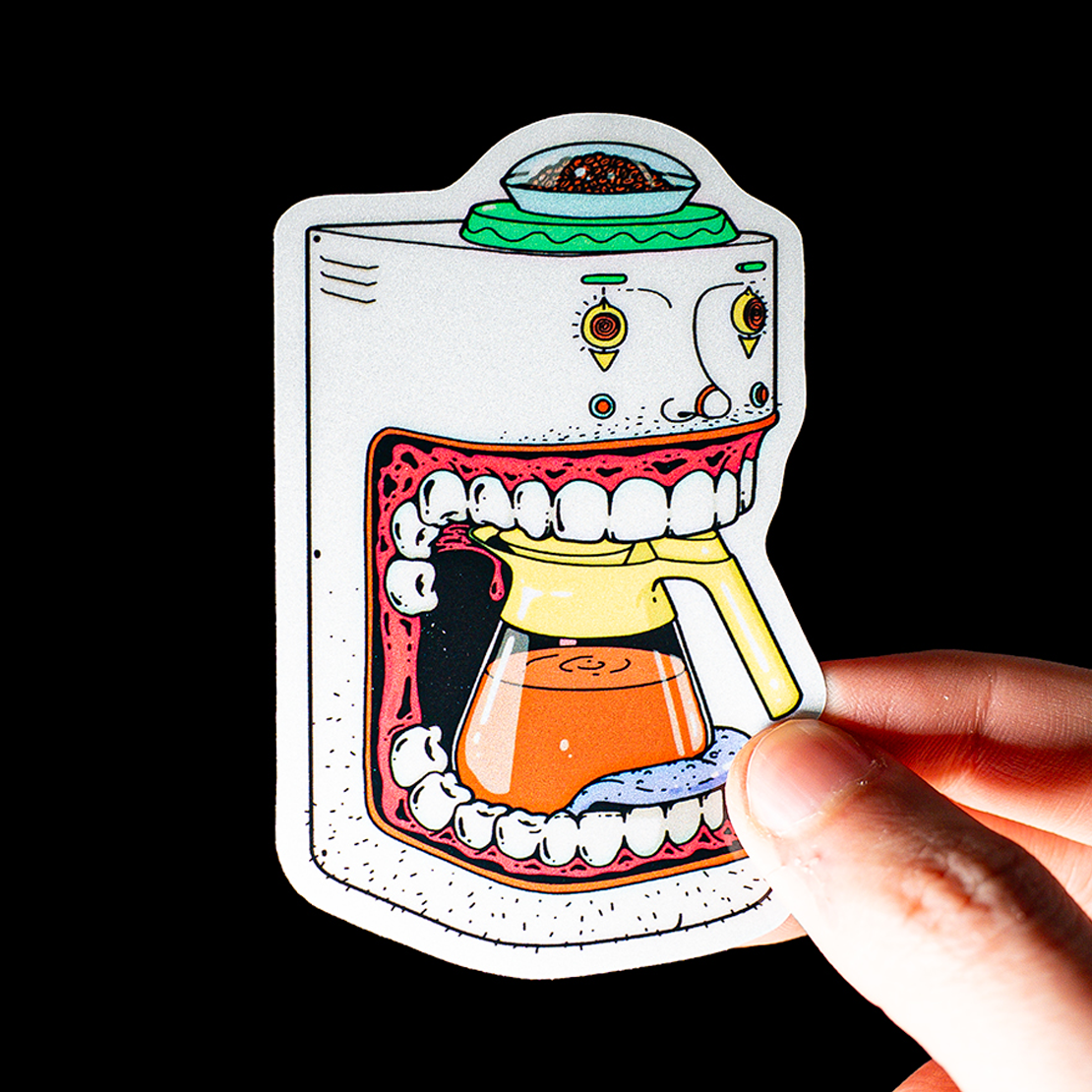 Cartoon sticker of a coffee maker with a mouth and teeth, holding a pot of coffee. A hand is holding the sticker against a black background.