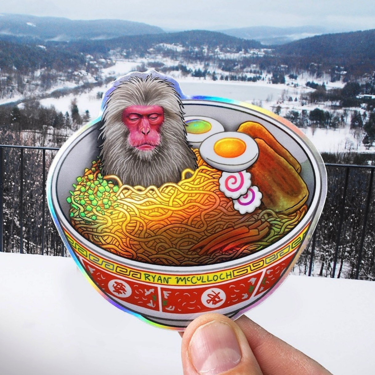 Die-cut holographic sticker of a monkey sitting in a ramen bowl with noodles, against a wintery background with mountains and snow