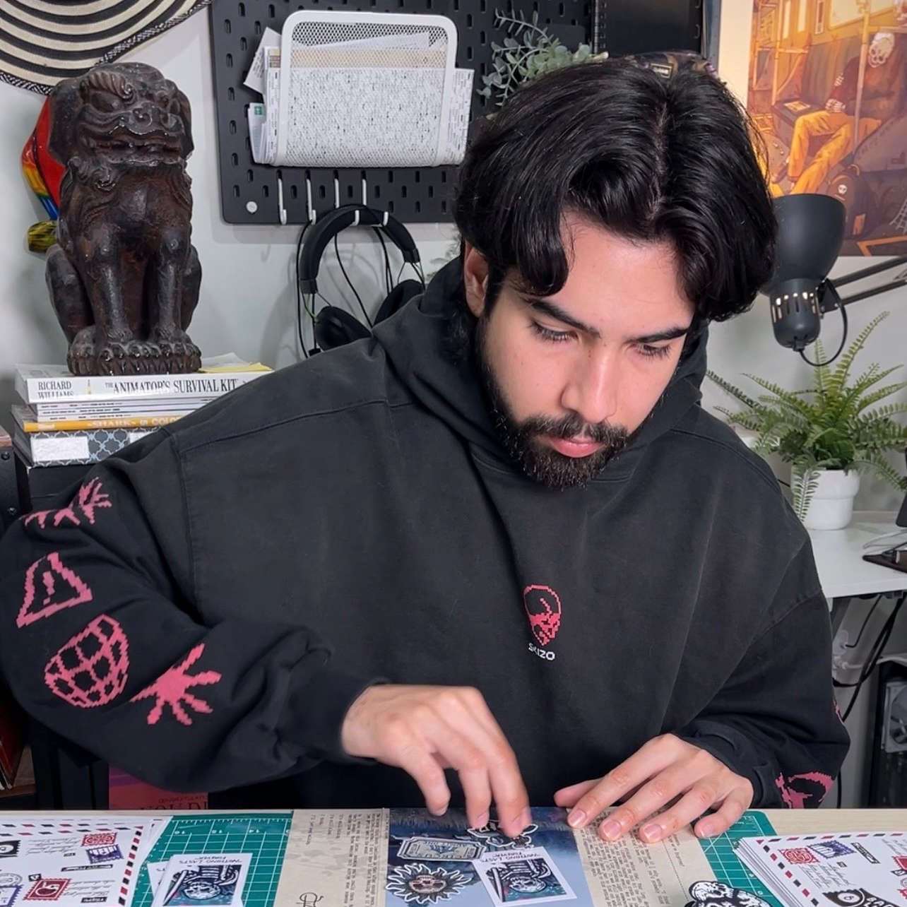 En man i svart hoodie sitter i sin studio och ordnar klistermärken på bordet framför sig, med skulpturer och konst i bakgrunden.