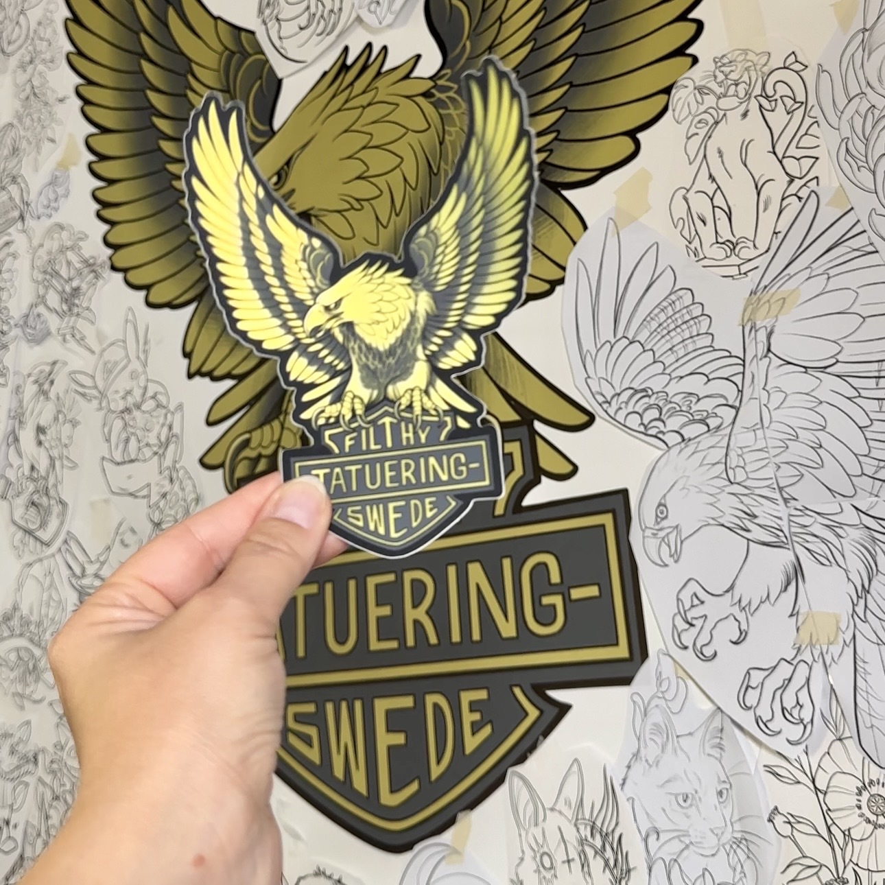 A matte mirror die-cut sticker held in front of a wall sticker with the same bird design, with the text "Filthy Swede Tatuering" in the middle, against a background of drawn tattoo designs.