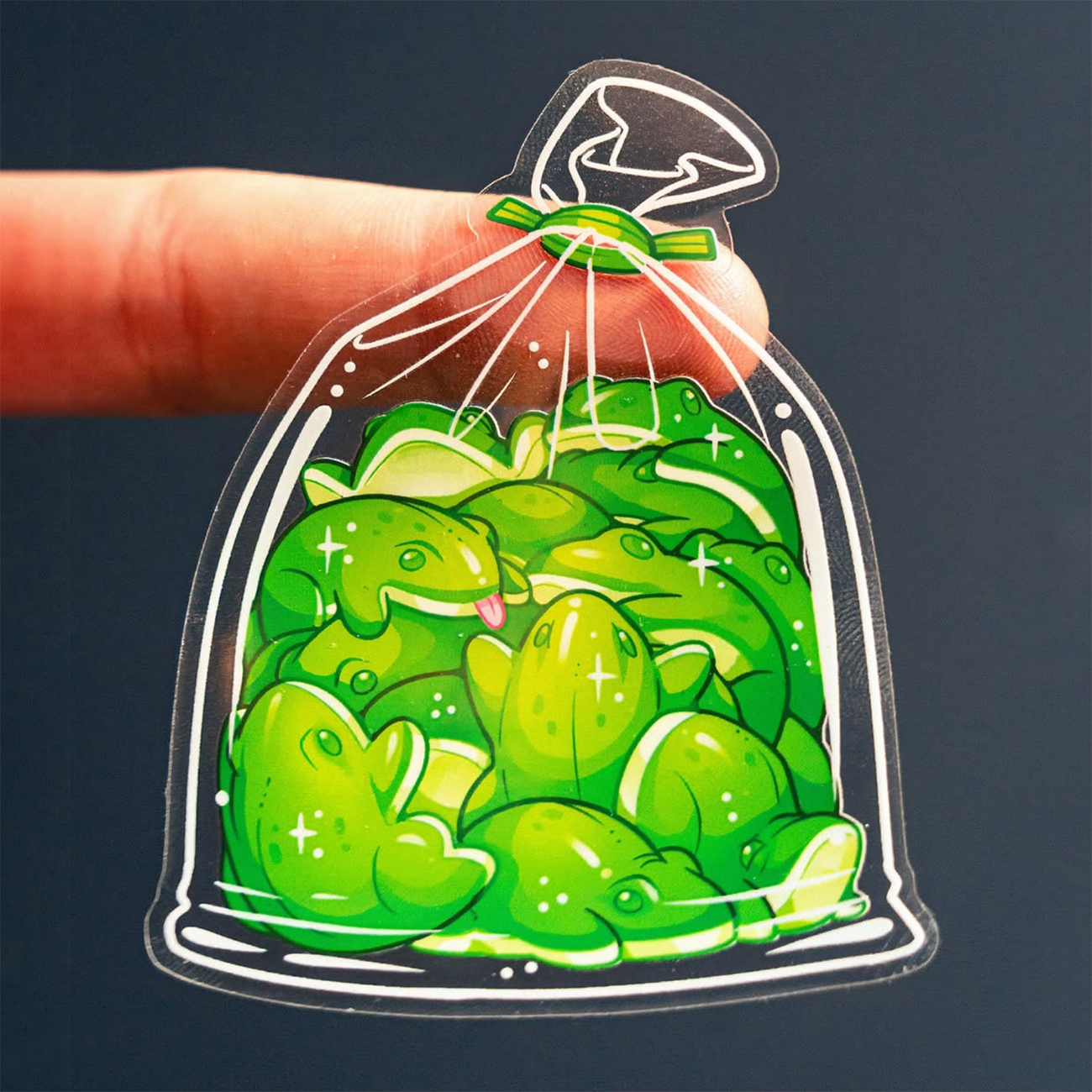 Finger holding frogs in a plastic bag, printed on transparent sticker material, held against a dark background.