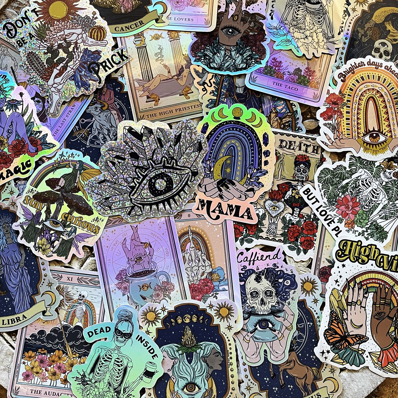 Collection of various colorful die-cut stickers laying on a table, in a spiritual, boho style, printed on glitter, holographic and vinyl.