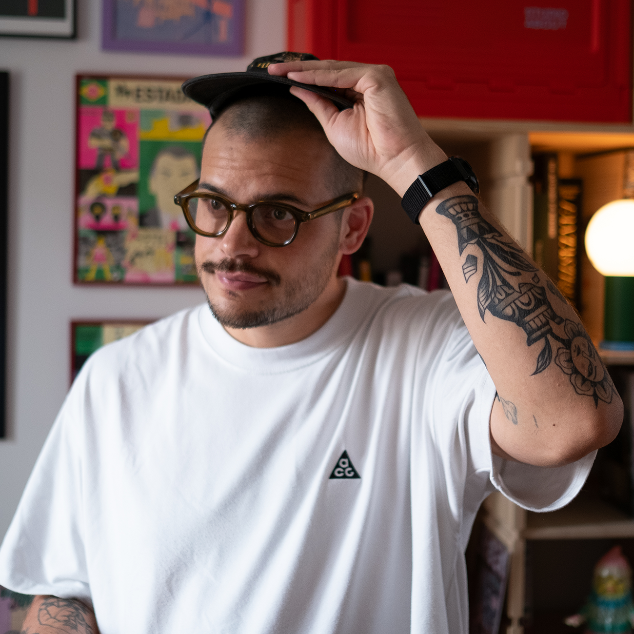 a man in white t-shirt holding his cap with one hand looking at something, with his studio as background