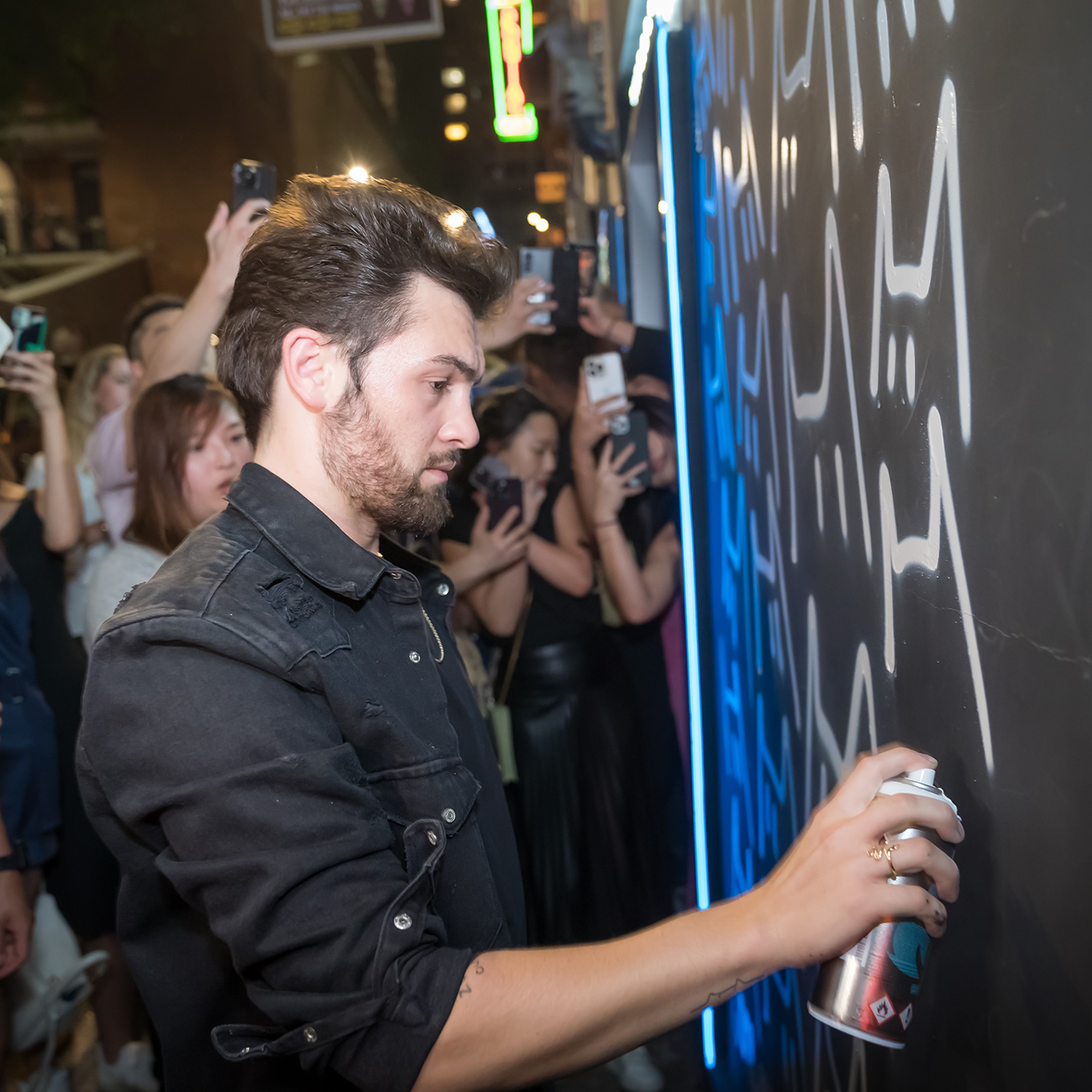 A man spray paints graffiti on a wall as a crowd watches and takes photos. He's wearing a black jacket.