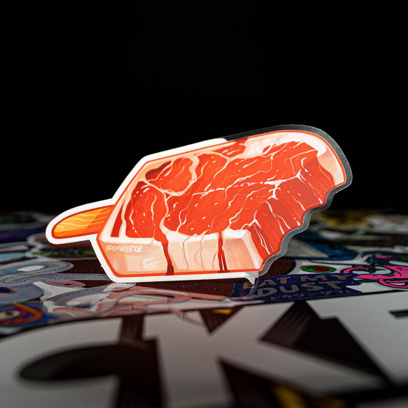 A steak formed as a popsicle, with a wooden handle and colorful artwork in the foreground against a black bakground.