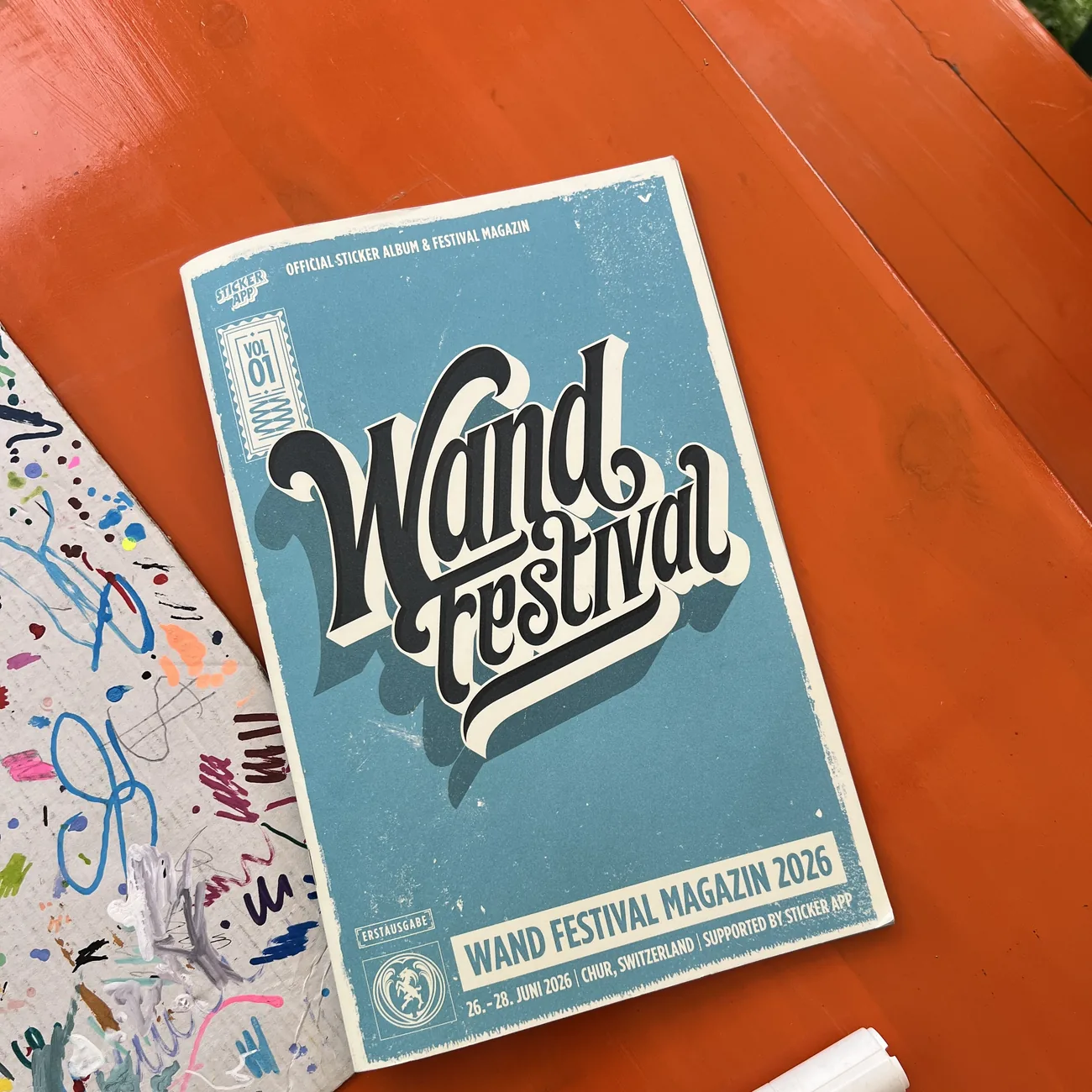 Wand Festivals officiella klistermärkesalbum och festivalmagasin 2026, med stöd av StickerApp, bredvid en färgpenna på ett orange bord
