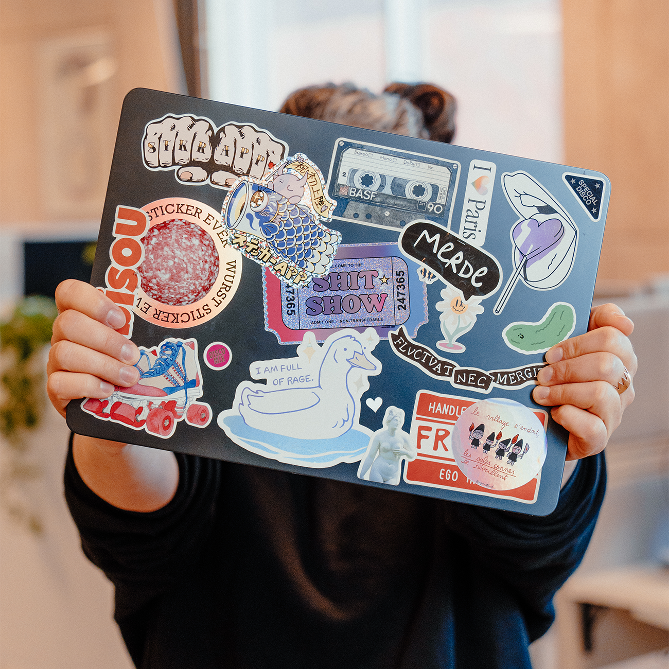 Närbild av en laptop täckt med en blandning av färgglada klistermärken, inklusive illustrerade karaktärer, typografi och grafiska designer, hålls upp av en person inomhus.
