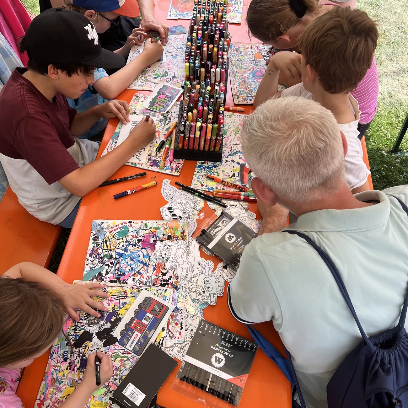 Barn färglägger die-cut klistermärken från Brainfart runt ett ställ med färgpennor på en festivalworkshop utomhus