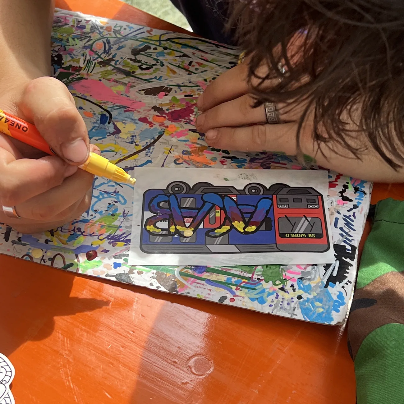 En hand färglägger en etikett med S8 World-tåget med gul färgpenna på workshoppen med Brainfart x StickerApp