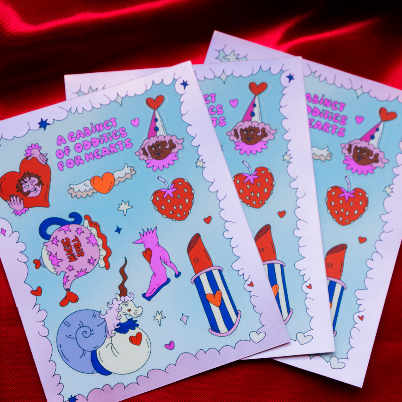 Pink sticker sheet for Valentine's Day with lipstick, snail, and a heart-shaped strawberry, against a silky red fabric.