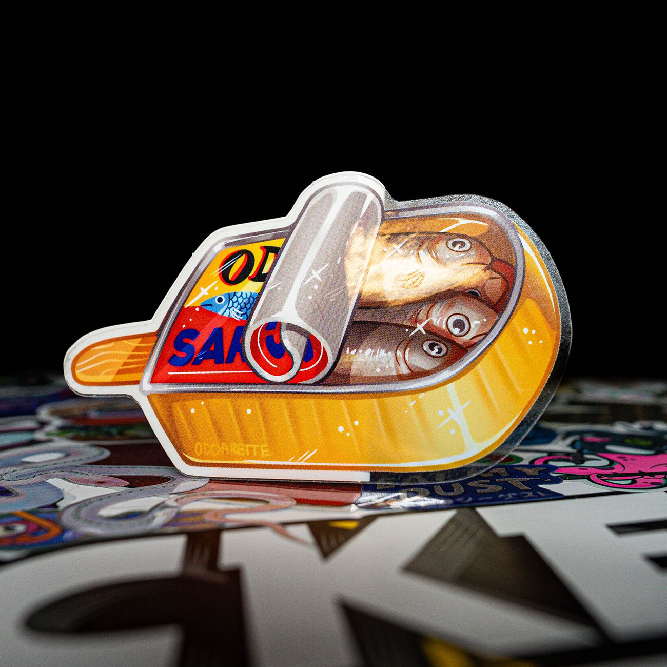 A sardine tin formed as a popsicle, with a wooden handle and colorful artwork in the foreground against a black bakground.