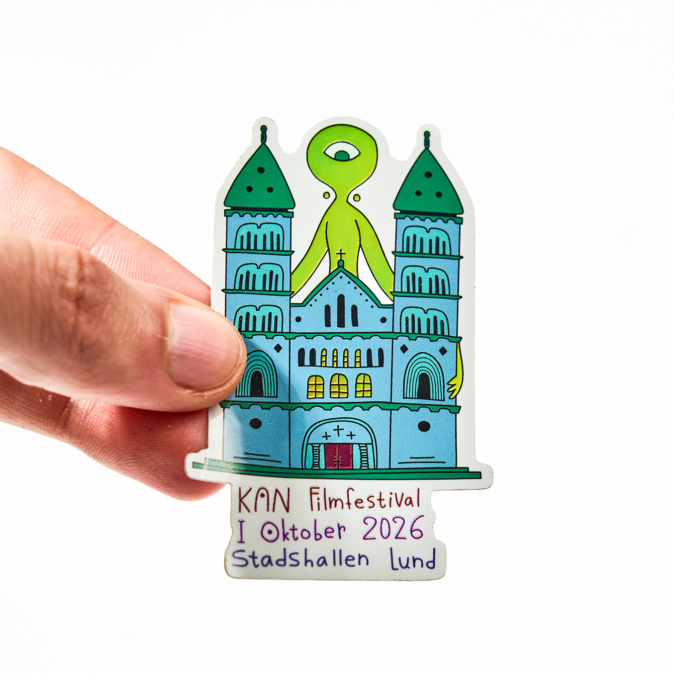 Hand som håller ett die-cut klistermärke för KAN Filmfestival, oktober 2026 i Stadshallen Lund, med en katedral och en grön enögd utomjording