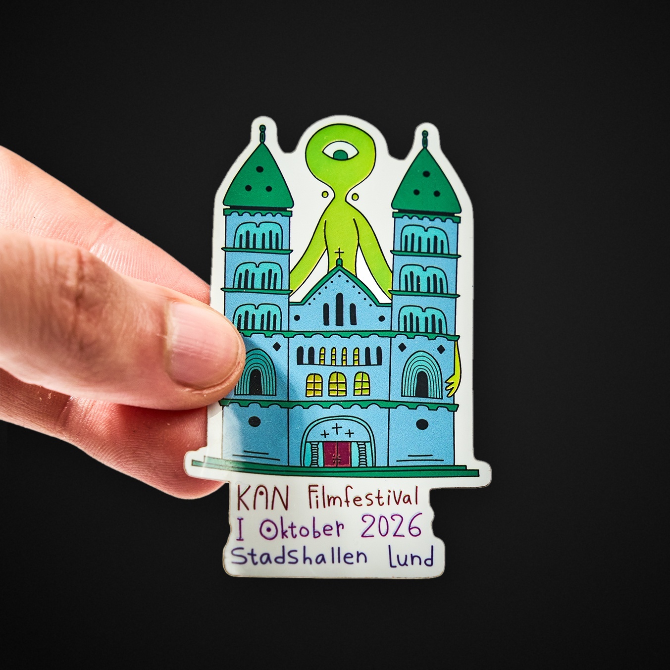 Hand som håller ett die-cut klistermärke föreställande en katedral med en grön enögd varelse och texten ”KAN Filmfestival I Oktober 2026 Stadshallen Lund”