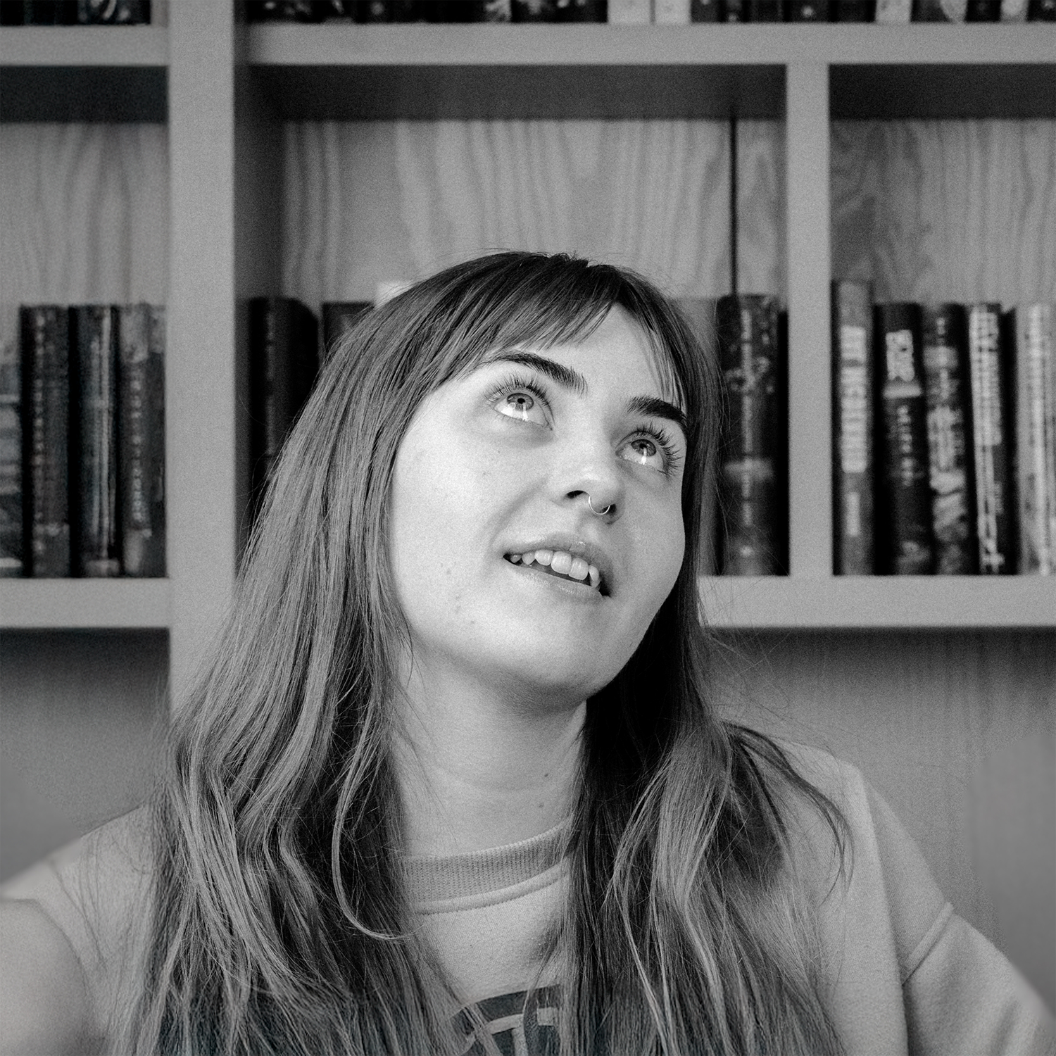 Woman with long hair looking upwards, sitting in front of a bookshelf filled with books. Black and white photo.