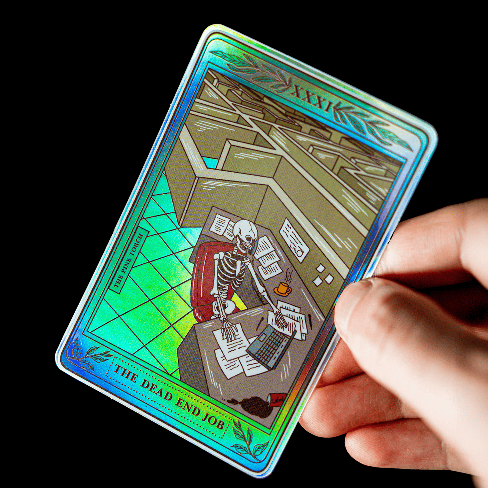 Hand som håller ett holografiskt tarotkortsklistermärke med titeln ”The Dead End Job” och ett skelett vid ett kontorsskrivbord