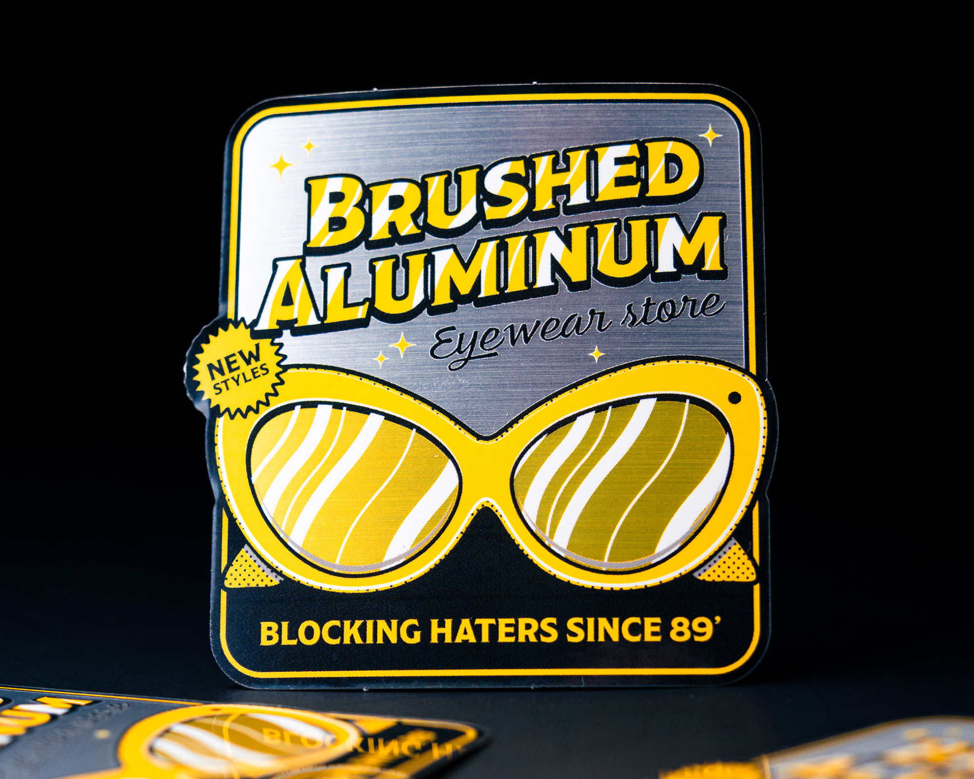 Retro-style sticker for "Brushed Aluminum Eyewear Store" featuring yellow sunglasses and the tagline "Blocking Haters Since 89'."