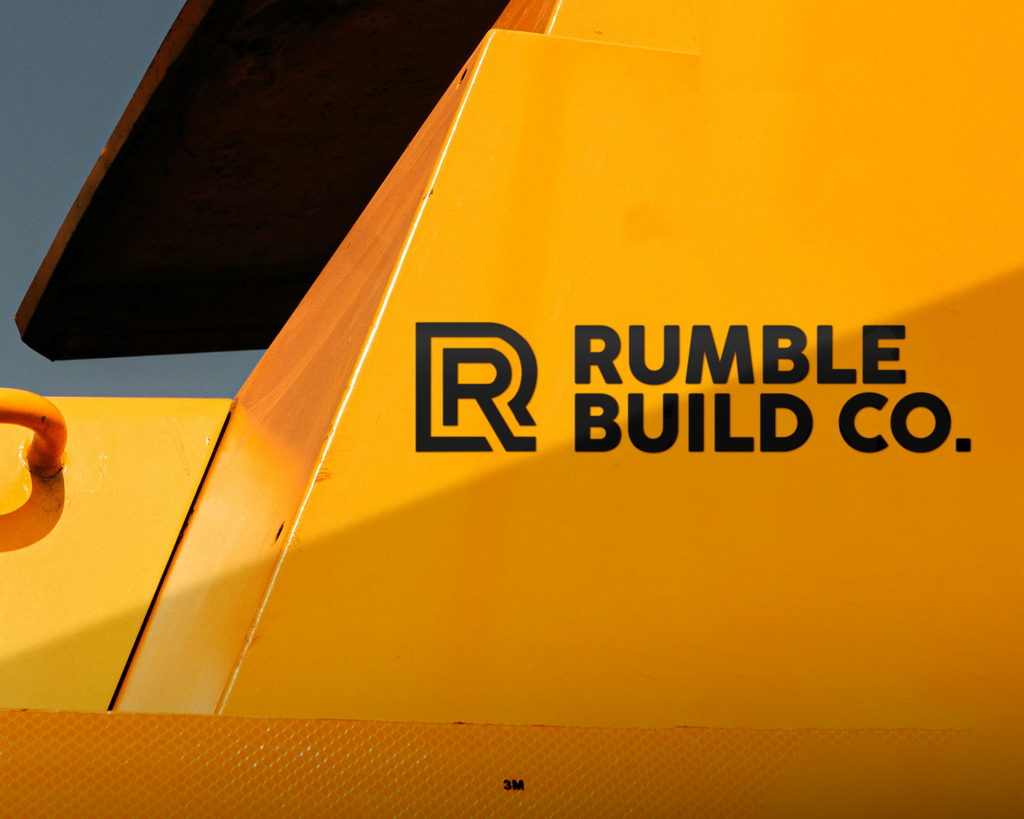 Närbild av ett gult industrifordon med "Rumble Build Co." vinyltransfer i fet stil på dess yta, med en abstrakt "R"-design.