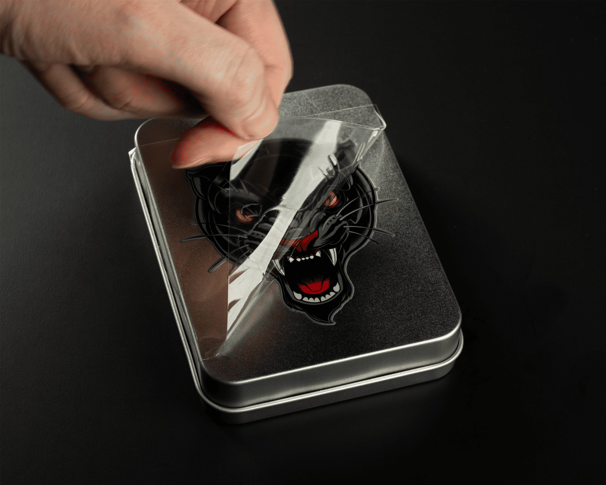 A hand peeling a protective film off a metal tin with a fierce panther design on the lid, set against a black background.