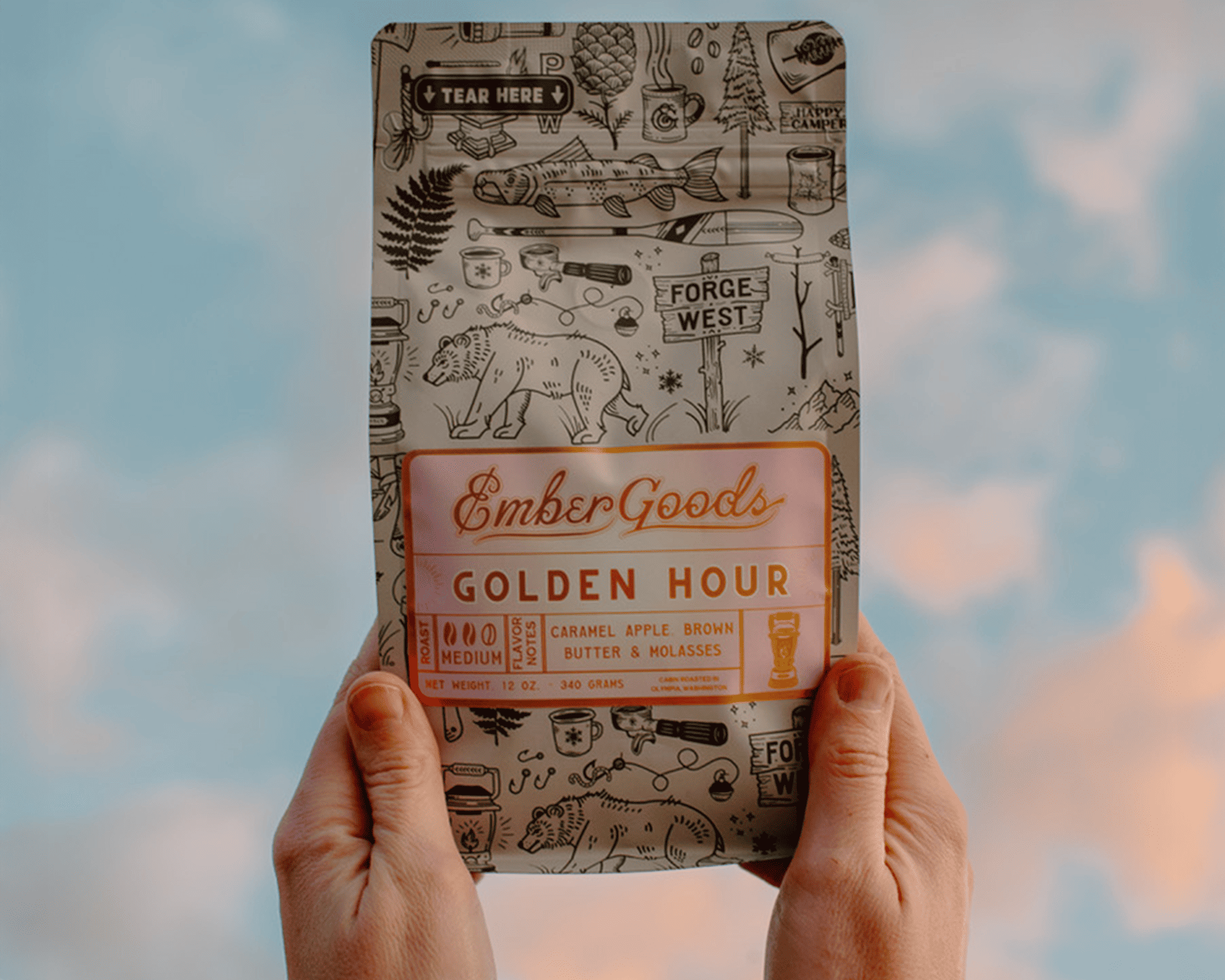 Händer som håller en påse Ember Goods Golden Hour-kaffe med metallisk etikett mot en himmel i bakgrunden