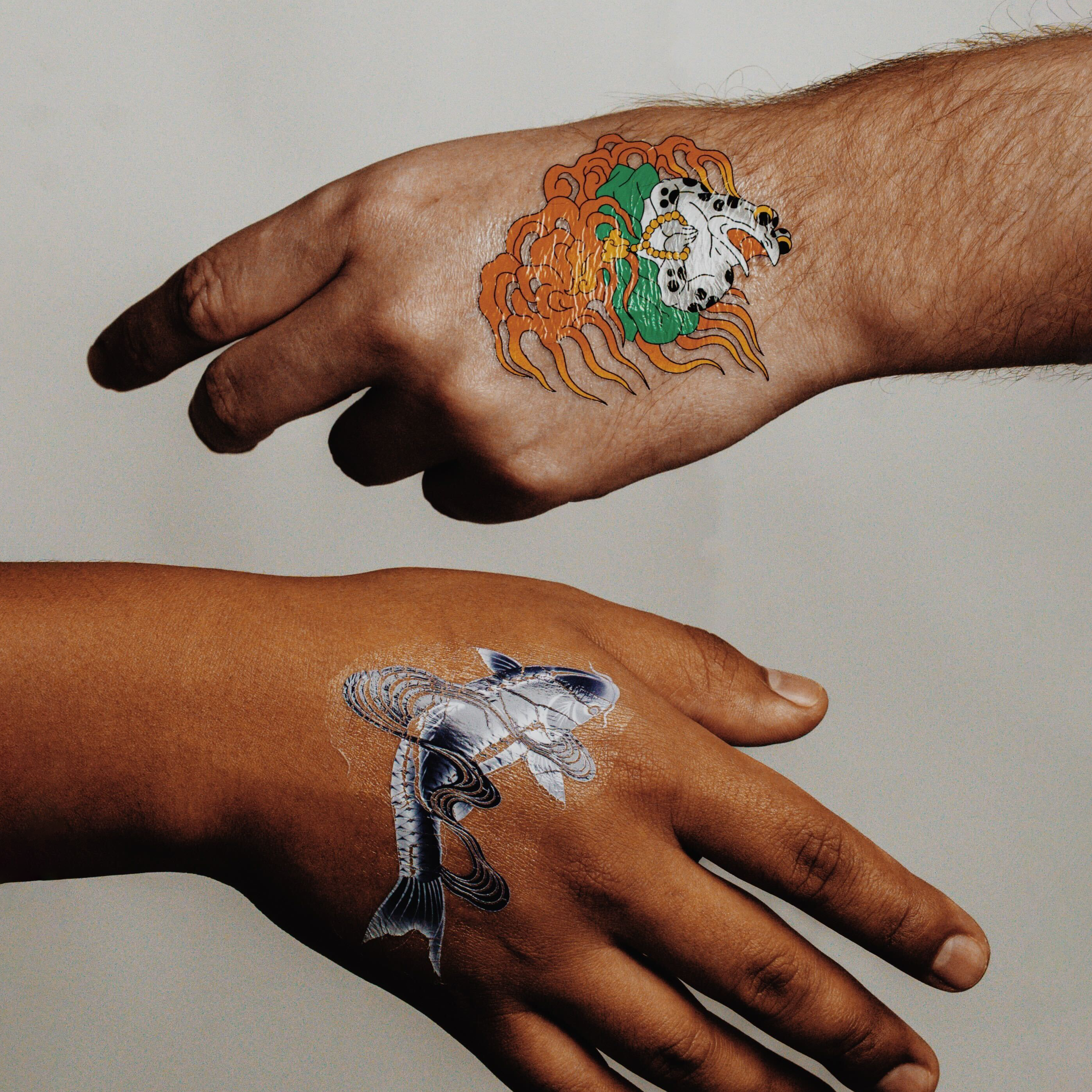 Temporary tattoos on two hands, one featuring a detailed koi fish design and the other a stylized skull with flowing orange elements, set against a neutral background.