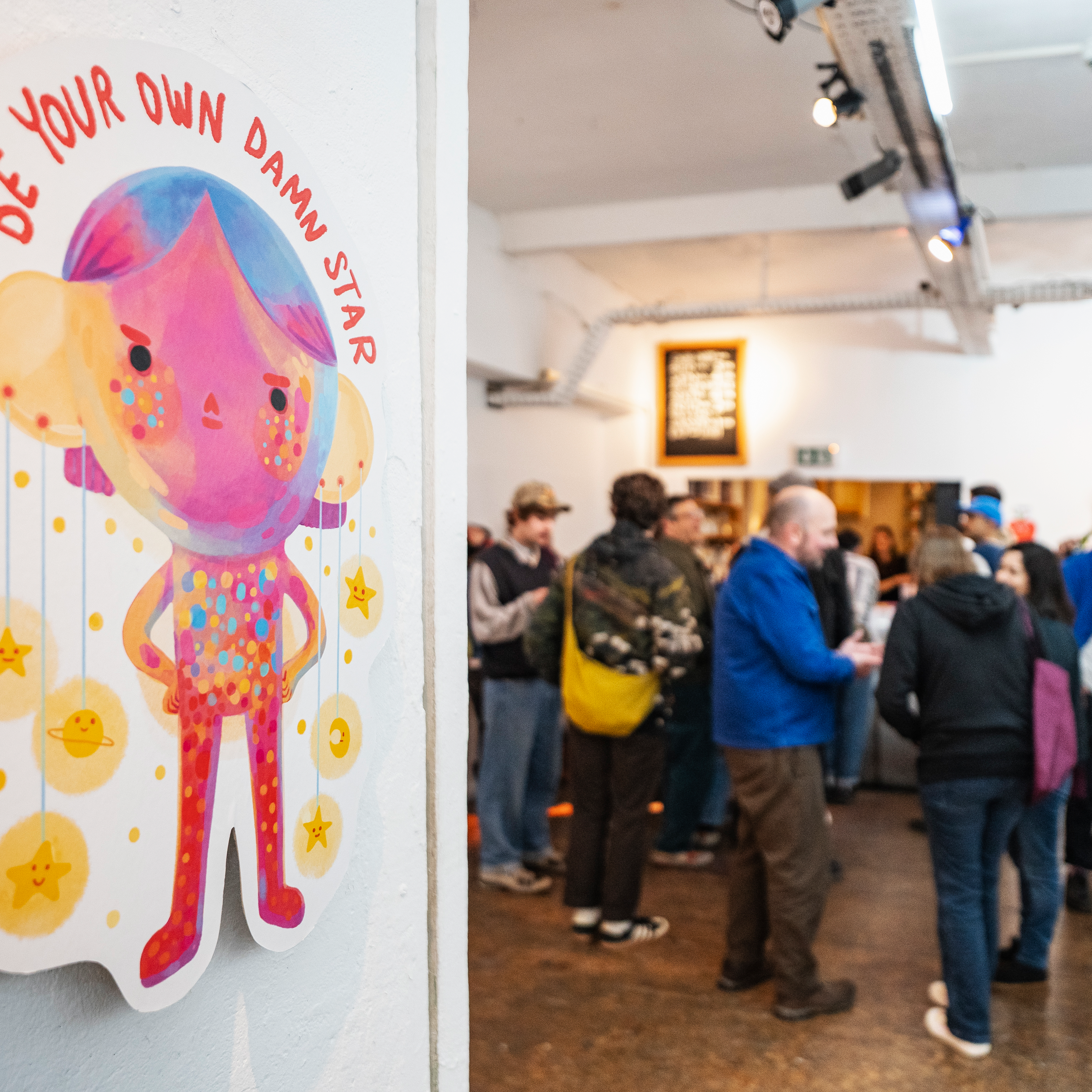 Visitors at an art exhibition talking, in the foreground is a piece of art hanging with the text "Be your own damn star".