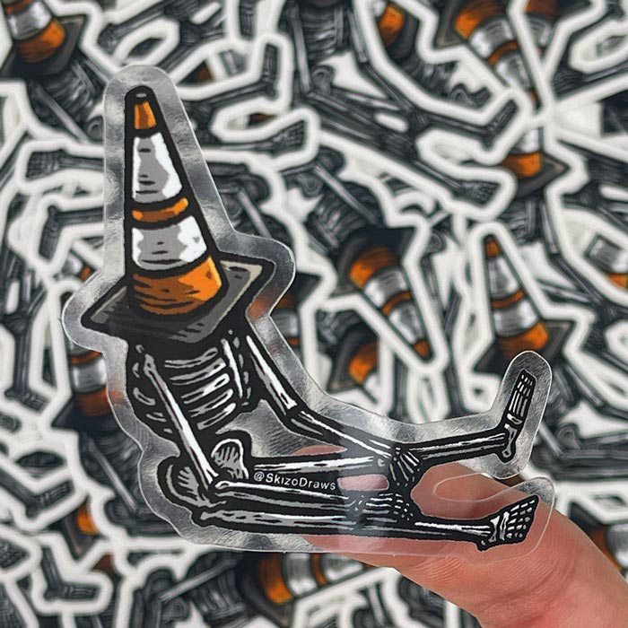 Transparent die-cut klistermärke av ett skelett som sitter på marken med en trafikkon på huvudet, mot en bakgrund av klistermärken.
