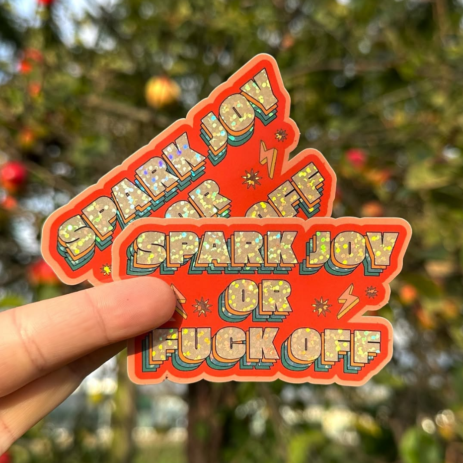 Glitter orange retro sticker with the text "spark joy or fuck off" held in front of an apple tree.